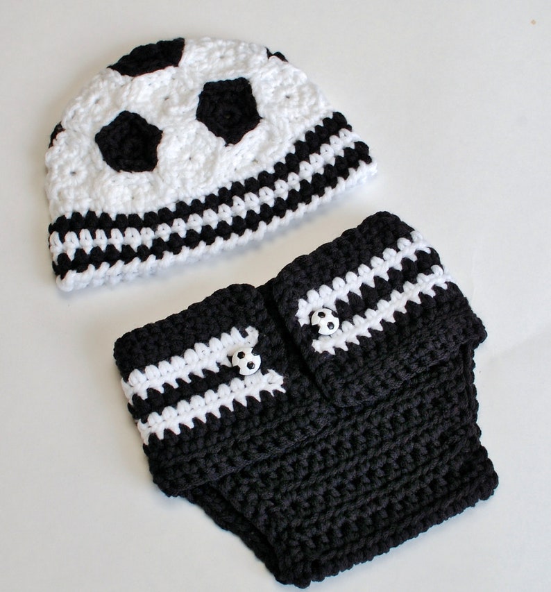 May include: A crocheted baby set featuring a soccer ball design. The set includes a white and black hat with soccer ball patches and a matching diaper cover. The diaper cover has white stripes and soccer ball buttons.