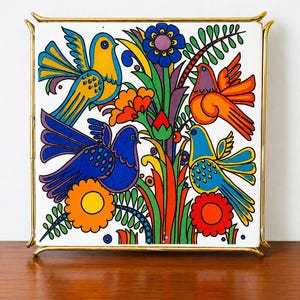 Op de afbeelding: Een vierkante keramische tegel met een levendige, veelkleurige illustratie van vier vogels omringd door gestileerde bloemen en bladeren, geplaatst in een dun, gepolijst goudkleurig metalen frame dat de tegel stevig op zijn plek houdt.