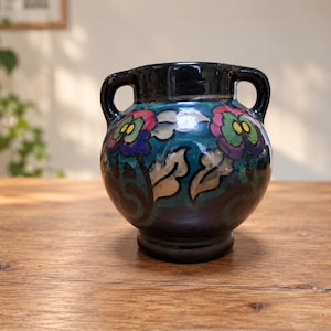 Może przedstawiać: Ceramiczny wazon z dwoma uchwytami, wykończony ciemnoniebieską i czarną glazurą. Korpus ozdobiony jest ręcznie malowanym wzorem kwiatowym w odcieniach różu, zieleni i bieli, przedstawiającym stylizowane kwiaty i liście na ciemnym tle.