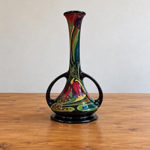 Op de afbeelding: Een keramieken vaas met een zwarte voet en twee handvatten, voorzien van een kleurrijk beschilderd ontwerp van rode en paarse tulpachtige bloemen, groene bladeren en gouden accenten. De hoge, slanke hals loopt uit naar de bovenkant.