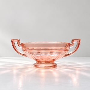 Op de afbeelding: Een doorschijnende roze glazen kom met twee symmetrische handvatten aan weerszijden, een ronde voet en een gefacetteerd patroon aan de buitenkant dat het licht opvangt en een stralende gloed op het oppervlak creëert.