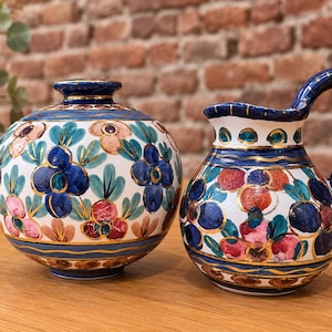 Peut inclure: Un vase en céramique rond et une carafe assortie, tous deux dotés d'une base blanche avec des motifs floraux peints à la main dans des tons de bleu, rose et vert, rehaussés de bordures dorées sur les cols, les anses et les bases.