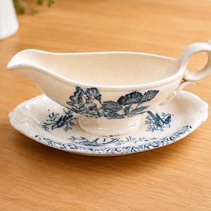 Op de afbeelding: Een crèmekleurige keramische sauskom met bijpassende ovale schotel, voorzien van een blauw bloemen- en vogelpatroon. De sauskom heeft een gebogen handvat, een brede schenktuit en een voetstuk, terwijl de schotel een geschulpte rand heeft.