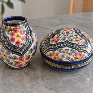 Puede incluir: Un jarrón de cerámica y una caja redonda a juego con tapa, ambos con una base de color crema y motivos florales pintados a mano en rosa, amarillo y morado, acentuados por bordes geométricos azul intenso y finas líneas de color dorado.