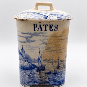 Op de afbeelding: Een rechthoekige keramische voorraadpot met deksel en handvat, voorzien van een blauw-witte transferware landschapsscène van een haven met zeilboten en een kerk. Het woord PATES staat in blauw gedrukt op de voorzijde van de gebroken witte pot.
