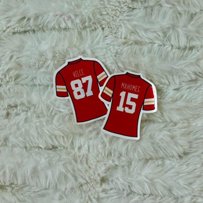 May include: Two red football jersey stickers. One sticker has the name "Kelce" and the number "87" in white. The other sticker has the name "Mahomes" and the number "15" in white. Both jerseys have white and gold stripes on the sleeves.