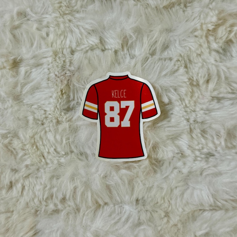 May include: A red sticker in the shape of a football jersey. The jersey has white numbers and text that read "87" and "Kelce". The jersey has white and yellow stripes on the sleeves.