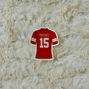 May include: A red sticker in the shape of a football jersey. The jersey has white and gold accents on the sleeves and shoulders. The name "Mahomes" and the number "15" are printed on the back of the jersey.