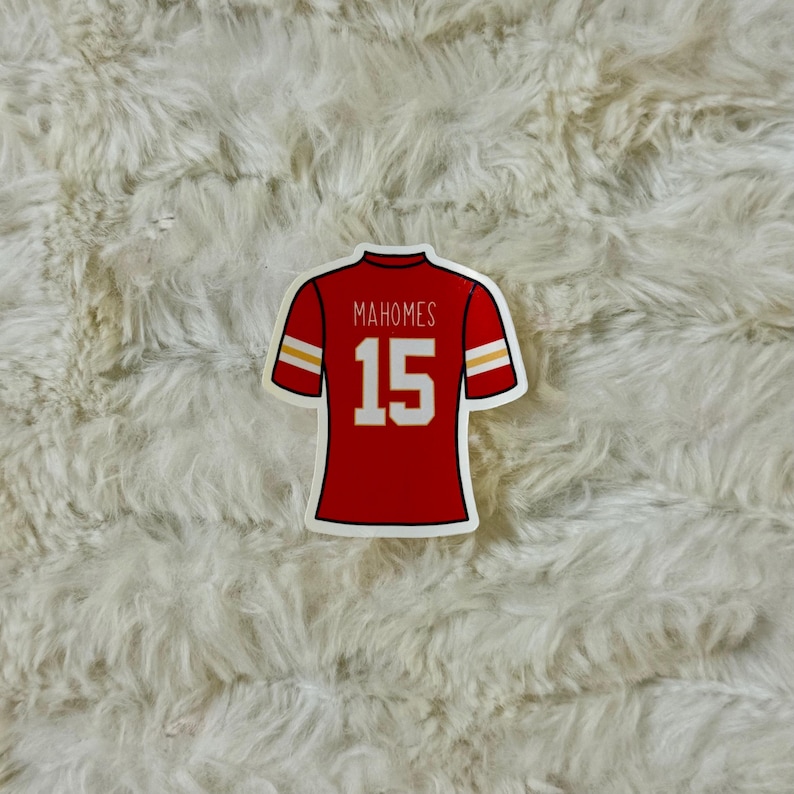 May include: A red sticker in the shape of a football jersey. The jersey has white and gold accents on the sleeves and shoulders. The name "Mahomes" and the number "15" are printed on the back of the jersey.