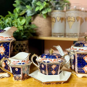 May include: A porcelain tea set featuring a tall coffee pot, a creamer, a lidded sugar bowl on a saucer, and a teapot. Each piece has a deep blue base with orange floral diamond patterns, gold trim, and white accents on a hexagonal body.