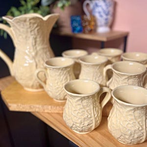 May include: A cream-colored ceramic pitcher and seven matching mugs, each featuring a raised, textured leaf pattern across the exterior. The pitcher has a wide, scalloped rim and a curved handle, while the mugs have rounded bodies.