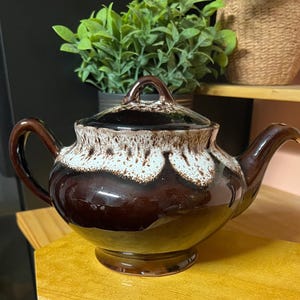 May include: A ceramic teapot featuring a glossy dark brown glaze on the lower body and a contrasting white drip glaze pattern around the upper rim and lid. The teapot has a curved handle, a tapered spout, and a small knob on the lid.
