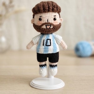 May include: A handcrafted crochet doll featuring a figure with brown hair and a beard, wearing a white and light blue striped jersey with the number 10 printed in black on the chest, paired with black shorts and white socks with light blue stripes.