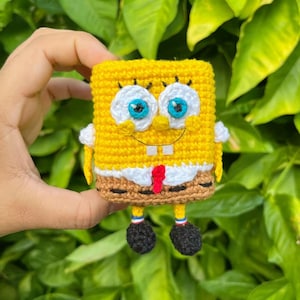 May include: A yellow, rectangular crochet figure resembling a sponge character with large white eyes, blue pupils, a small yellow nose, and a wide smile. It wears a white shirt with a red tie and brown pants, featuring small black shoes and arms.