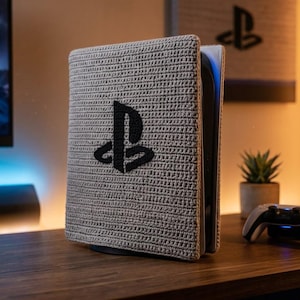 Könnte beinhalten: Eine aufrecht stehende Spielkonsole, die mit einer hellgrauen, strukturierten Häkelhülle bedeckt ist. Auf der Vorderseite der Hülle befindet sich ein markantes, schwarz gesticktes PlayStation-Logo. Die Konsole steht neben einem weißen Controller.