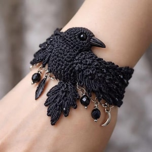 May include: A black crochet bracelet featuring a detailed bird design with a smooth beak and a round eye. The piece is adorned with dangling silver-toned charms, including small black faceted beads, a feather shape, and a crescent moon charm.