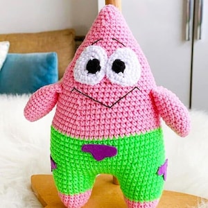 Puede incluir: Un peluche de Patrick Estrella hecho a mano con ganchillo. El juguete presenta un cuerpo rosa, pantalones cortos verdes con detalles morados y ojos blancos grandes con pupilas negras. Los brazos y las piernas también son rosas. El juguete está sobre una superficie de madera.