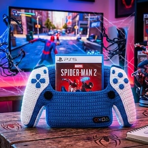 May include: A blue and white crocheted controller holder shaped like a game controller, featuring a central slot holding a Marvel Spider-Man 2 game case for the PS5. The holder has a small black tag with white geometric symbols on the front right side.