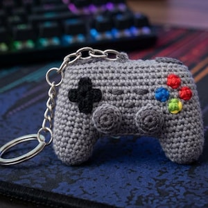 May include: A grey crocheted video game controller keyring featuring a black directional pad, two circular grey thumbsticks, and four small buttons in red, blue, green, and yellow. The controller is attached to a silver metal split ring.