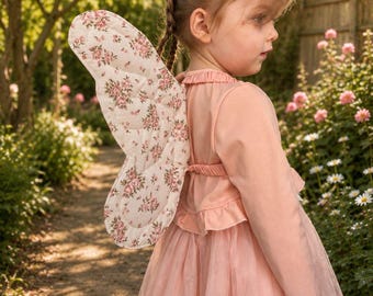 May include: A light pink long-sleeved dress with a ruffled bodice and a layered tulle skirt, paired with quilted fabric wings featuring a pink and green floral pattern on a cream background, worn by a child with braided hair in a garden setting.