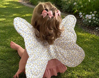 May include: A pair of quilted butterfly wings featuring a light blue and white floral pattern. The wings are worn over a dusty pink ruffled top. The hair is styled with two small pink bows, and the wings are shaped like large insect wings.