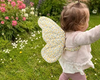 May include: A pair of quilted butterfly wings featuring a yellow and white floral pattern on a white background, attached with matching fabric straps. The wings are worn over a light pink long-sleeved top with a white lace-trimmed hem and pink pants.