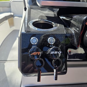 May include: A black boat dashboard console featuring a circular cup holder, two silver push-button switches labeled HORN and BILGE, and two black circular knobs with the text AR and 220, each attached to a small black plastic tethered key.
