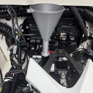 May include: A gray 3D-printed funnel with a wide conical top and a narrow cylindrical stem, positioned vertically within a mechanical engine compartment. The funnel features a small side tab near the rim and a textured surface finish.