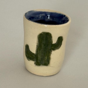 Pode incluir: Uma pequena chávena de cerâmica cilíndrica com um exterior branco sujo e um interior azul profundo. A superfície exterior apresenta um design de cato verde escuro pintado à mão com dois braços no tronco central.