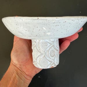 Op de afbeelding: Een witte keramische voetbakschaal met een gespikkeld oppervlak. De kom wordt ondersteund door een cilindrische basis met een gestructureerd, patroonontwerp. De kom wordt in een hand gehouden tegen een donkere achtergrond.