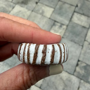 Puede incluir: Un anillo de cerámica grueso y cilíndrico con un patrón repetitivo de rayas verticales de color marrón texturizado que alternan con rayas blancas lisas y brillantes. La superficie de las secciones marrones es granulada, mientras que las blancas están esmaltadas.