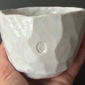 Könnte beinhalten: Eine weiße Keramikschale mit einer strukturierten, facettierten Außenseite. Die Schale hat ein kleines, kreisförmiges Emblem in der Nähe des Bodens. Die Schale wird in einer Hand gehalten. Die Schale ist wahrscheinlich handgefertigt und zum Servieren von Speisen geeignet.