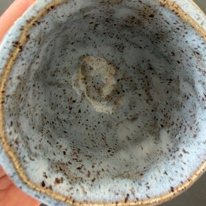 Puede incluir: Una taza de cerámica con esmalte azul claro y motas de color marrón oscuro en el interior. El borde de la taza presenta un acabado texturizado de tono metálico que contrasta con la superficie azul moteada de las paredes interiores.