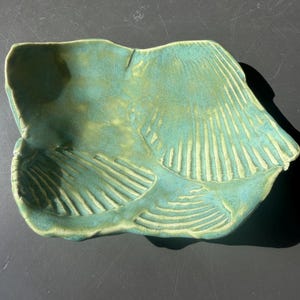 May include: A hand-formed ceramic dish with an irregular, organic shape and a glossy teal and light green glaze. The surface features embossed, fan-shaped shell patterns with raised ridges, creating a textured, multi-dimensional appearance.