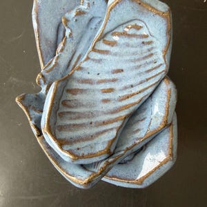 Puede incluir: Una pila de tres platos de cerámica de forma irregular hechos a mano, con un esmalte azul claro brillante y bordes de arcilla marrón expuestos. Cada plato presenta una superficie texturizada con estrías horizontales y una forma orgánica.