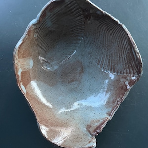May include: A hand-formed ceramic bowl with an irregular, organic shape resembling a seashell. The interior features a glossy, speckled light gray glaze with visible ridges and textures, while the outer edges show a warm, earthy brown tone.