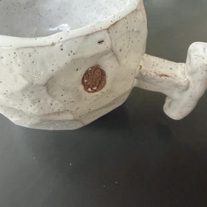 Puede incluir: Taza de cerámica blanca hecha a mano con superficie texturizada y asa única. La taza tiene un aspecto moteado y un pequeño emblema circular marrón. Ideal para bebidas calientes.