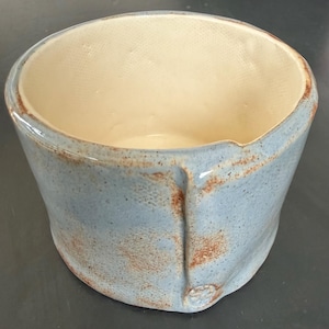 Puede incluir: Un cuenco de cerámica hecho a mano con un esmalte azul claro moteado y un interior liso de color crema. El exterior presenta un detalle de cresta vertical y manchas de óxido de hierro marrón rojizo, lo que le da un aspecto texturizado y orgánico.