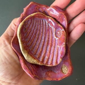 Könnte beinhalten: Eine kleine, handgeformte Keramikschale mit einem muschelförmigen Zentrum und erhabenen Rippen, glasiert in leuchtenden Magenta-, Violett- und Orangetönen. Das Stück hat einen gesprenkelten, unglasierten beigen Rand und einen kleinen kreisförmigen Stempel.