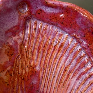 Puede incluir: Un cuenco de cerámica con un esmalte brillante y moteado en tonos magenta vibrante, rojo intenso y naranja. La superficie interior presenta una serie de crestas o surcos verticales en relieve que recorren la base curva con textura.