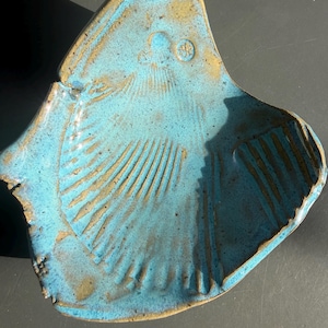May include: A ceramic dish shaped like a scallop shell with a vibrant turquoise blue glaze. The surface features raised, radiating ridges and a small circular maker's mark near the top edge. The edges show exposed brown clay.