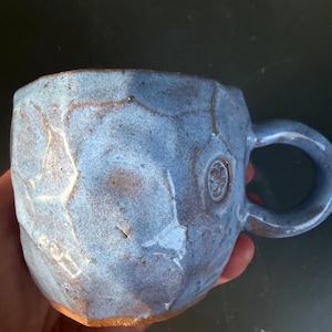 Puede incluir: Una taza de cerámica hecha a mano con una superficie texturizada y facetada y un esmalte azul. La taza tiene un asa redondeada y un pequeño diseño en relieve cerca del asa. La base de la taza tiene un acabado natural sin esmaltar.
