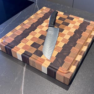 Può includere: Un tagliere quadrato in legno con un motivo a scacchiera multicolore nei toni del marrone scuro, marrone chiaro e crema. Il tagliere presenta un bordo scanalato e un coltello da cucina in acciaio inossidabile con manico nero appoggiato sopra.