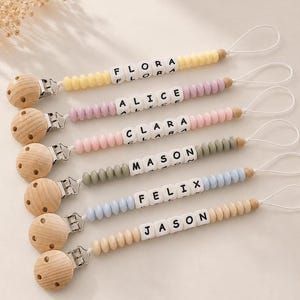 May include: Six personalised dummy clips featuring round wooden clips, white letter beads spelling FLORA, ALICE, CLARA, MASON, FELIX, and JASON, and colourful silicone beads in yellow, lavender, pink, sage green, light blue, and beige.
