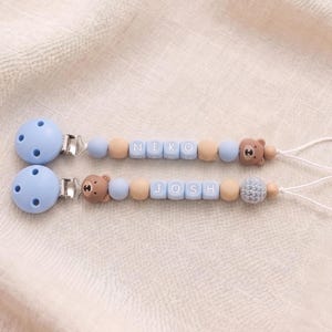 May include: Two dummy clips featuring light blue circular clips, silicone beads in shades of blue and tan, and bear-shaped accents. One clip displays the name NIKO on square beads, while the other displays the name JOSH on square beads, both ending with a white loop.