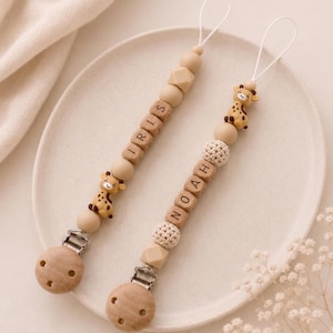 Könnte beinhalten: Zwei Schnullerketten mit Holzperlen, Giraffen-Anhängern aus Silikon und runden Holzclips. Eine Kette ist mit dem Namen IRIS und die andere mit dem Namen NOAH personalisiert, beide verfügen über weiße Schlaufen zur Befestigung an Kleidung.