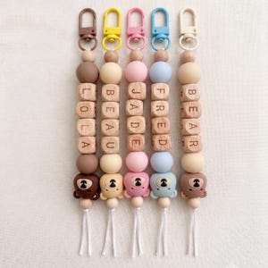May include: Five vertical keychains featuring silicone and wooden beads in brown, yellow, pink, blue, and beige. Each keychain has a plastic clasp, four wooden letter beads spelling names like LOLA, BEAU, JADE, FRED, and BEAR, and a bear-shaped charm.