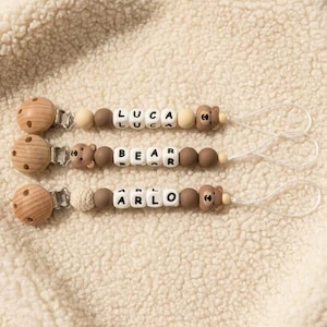 May include: Three dummy clips featuring wooden circular clips, silicone beads in shades of brown and cream, and bear-shaped charms. The beads spell out the names LUCA, BEAR, and ARLO in black letters on white cubes, finished with white loops.