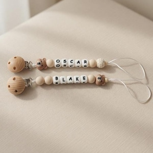 May include: Two dummy clips featuring round wooden clips, brown bear-shaped beads, and white letter beads spelling the names OSCAR and BLAKE. Each clip includes smooth wooden beads, a small crocheted bead, and a white cord loop for attachment.