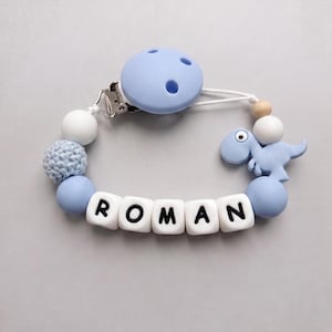 May include: A dummy clip featuring a light blue silicone dinosaur, white letter beads spelling ROMAN, light blue and white round silicone beads, a textured light blue crochet bead, and a light blue circular plastic clip with three ventilation holes.